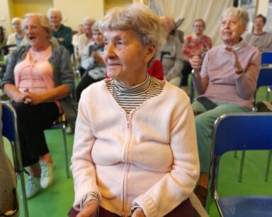 Sala. Uśmiechnięci seniorzy siedzą na krzesłach i klaszczą w dłonie. W tle kotary i duże piłki do ćwiczeń.