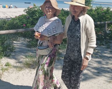 Zbliżenie. Dwie seniorki idą promenadą, trzymają się pod rękę. Na głowach mają kapelusze. W tle róże, piach, morze.