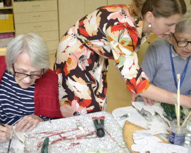 Zbliżenie. Młoda kobieta w kwiecistej sukience pomaga seniorkom w kolorowaniu roślinnych elementów, zieloną farbą.