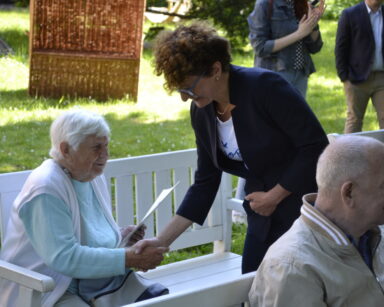 Ogród. Zbliżenie. Prezydentka Sopotu ściska dłoń seniorce, która siedzi na ławeczce i trzyma dyplom.