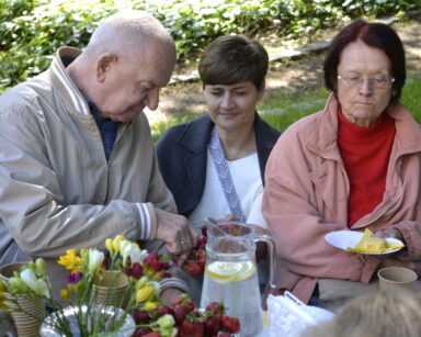 Zbliżenie. Kobieta kuca między parą seniorów, trzyma miskę truskawek. Senior wybiera truskawkę. Seniorka trzyma ciasto.