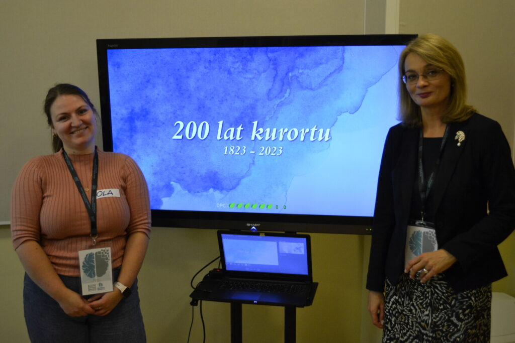 Zbliżenie. Dwie kobiety stoją przy monitorze z napisem "200 lat kurortu 1823-2023".