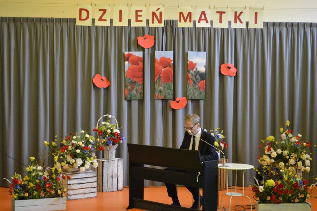Sala. Scena. Pianino, przy nim mężczyzna w garniturze i okularach. Obok kosze kwiatów, nad sceną napis" Dzień Matki".