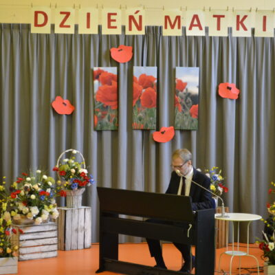 Sala. Scena. Pianino, przy nim mężczyzna w garniturze i okularach. Obok kosze kwiatów, nad sceną napis" Dzień Matki".