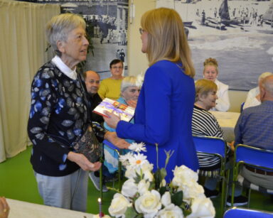 Sala. Kobieta w ciemnej marynarce wręcza praliny dla seniorki. W tle grupa seniorów przy stole. Na stole wazon z kwiatami.