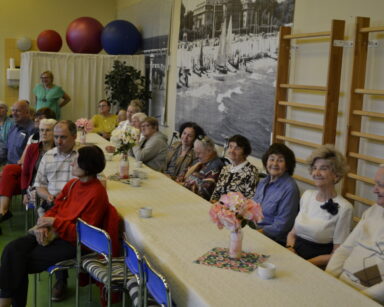 Sala. Grupa seniorów siedzi przy stole. Wszyscy uśmiechają się. Na stole filiżanki, wazony z kwiatami. W tle zdjęcia.