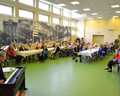 Sala. Mężczyzna przy pianinie. Na sali przy stołach siedzą uśmiechnięci seniorzy i opiekunowie. W tle na ścianie zdjęcia.