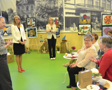 Sala. Trzy kobiety stoją przed grupą siedzących kobiet. Jedna z kobiet siedzących unosi rękę i coś mówi.