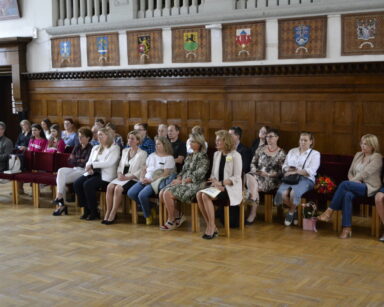 Sala. Grupa osób siedzi pod zdobioną drewnianą ścianą. Nad zdobieniami herby na arrasach i zdobiona balustrada balkonu.