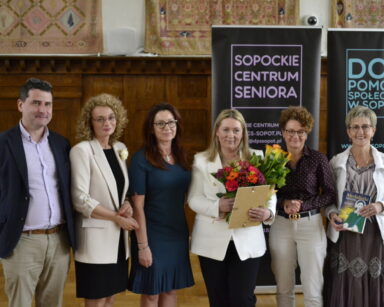 Zbliżenie. Sześć osób pozuje do pamiątkowego zdjęcia. Wszyscy odświętnie ubrani. W tle drewniana ozdobna ściana, arrasy.