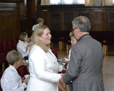 Zbliżenie. Mężczyzna w szarym garniturze składa gratulacje kobiecie w białej marynarce. W tle grupa ludzi.