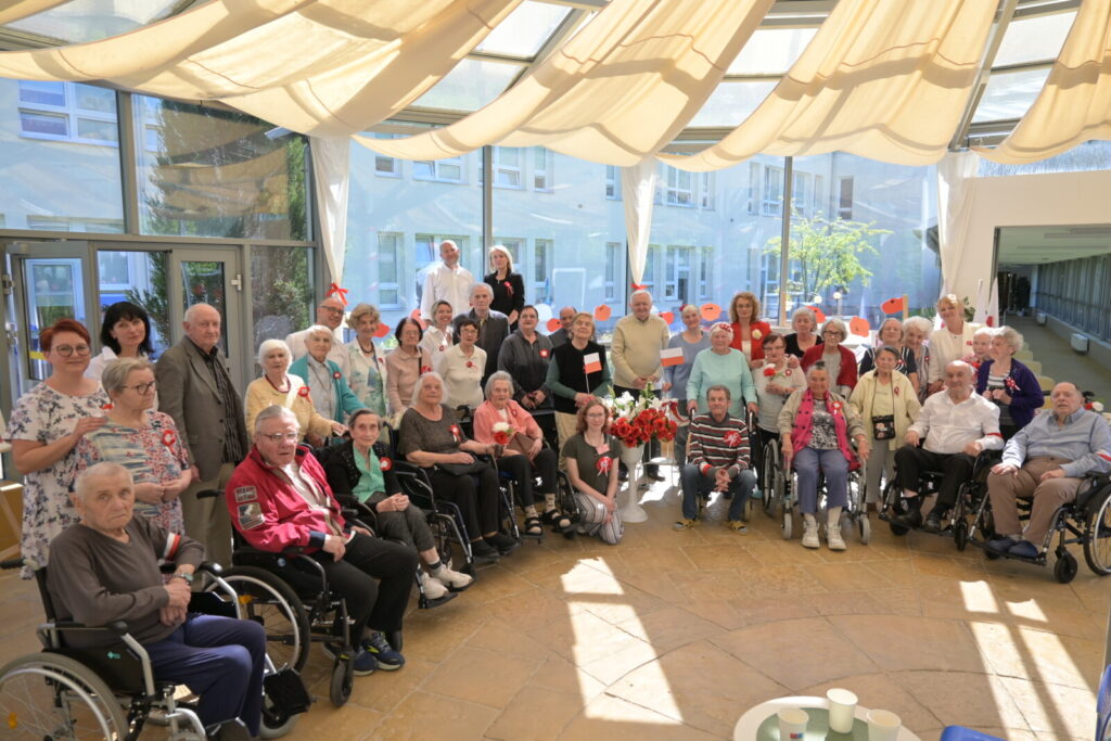 Sala. Grupa seniorów i opiekunów pozuje do zdjęcia. W rękach część osób trzyma flagi, część na piersi przypięty kotylion.