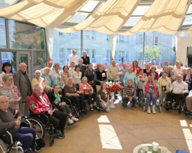 Sala. Grupa seniorów i opiekunów pozuje do zdjęcia. W rękach część osób trzyma flagi, część na piersi przypięty kotylion.