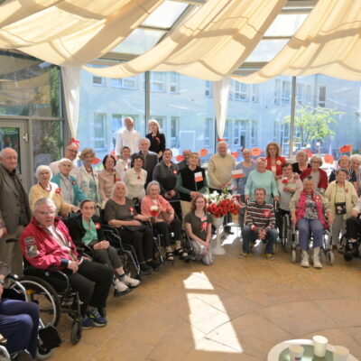 Sala. Grupa seniorów i opiekunów pozuje do zdjęcia. W rękach część osób trzyma flagi, część na piersi przypięty kotylion.