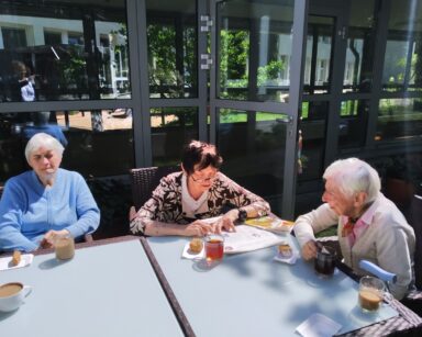 Zbliżenie. Troje seniorów siedzi przy stoliku. Na stole cztery kubki kawy, herbata, ciasto. Seniorka czytająca gazetę.