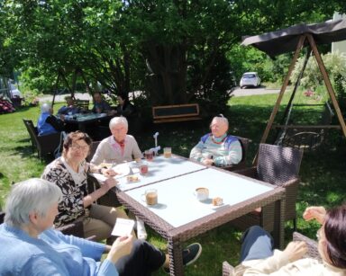 Ogród. Przy stolikach siedzą seniorzy. Piją kawę i herbatę, jedzą ciasto. Obok huśtawka z zadaszeniem i ławka pod drzewem.