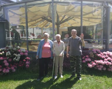 Ogród, zbliżenie. Troje seniorów pozuje do zdjęcia na tle ogrodu zimowego. Obok krzewy rododendronów i berberys.