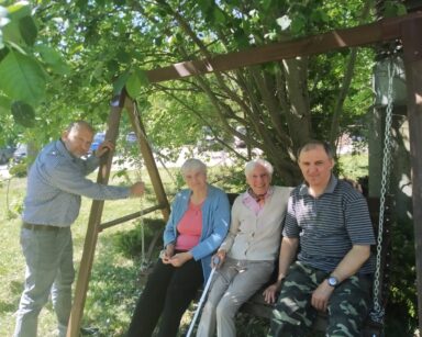 Ogród, zbliżenie. Troje seniorów siedzi na huśtawce z zadaszeniem. Obok stoi mężczyzna i trzyma rękę na łańcuchu huśtawki.