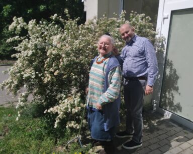 Zbliżenie. Seniorka i opiekun pozują do zdjęcia przy wielkim krzewie kwitnącym na biało. Oboje uśmiechają się.