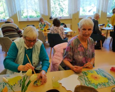 Sala. Seniorzy siedzą przy stołach i malują farbami wycięte z kartonu kwiaty słonecznika. Na stołach farby, słoiki z wodą.