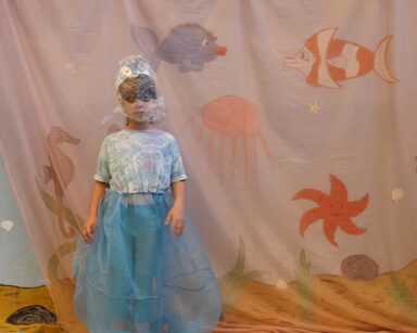 Zbliżenie. Na scenie dziewczynka ubrana w niebieską sukienkę, na głowie ma rybkę i worek foliowy. W tle brudne morze.