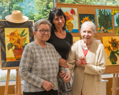Zbliżenie. Kobieta z dwiema uśmiechniętymi seniorkami pozuje do zdjęcia. W tle tablica z obrazami inspirowanymi Van Goghiem.