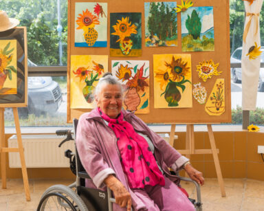 Zbliżenie. Uśmiechnięta seniorka na wózku w różowym dresie pozuje do zdjęcia. W tle obrazy inspirowane Van Goghiem.
