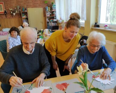 Sala. Młoda kobieta pochyla się nad seniorami, patrzy na ich prace. Seniorzy malują farbami wycięte kwiaty.