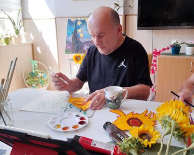 Sala. Stół. Senior maluje kwiat słonecznika. Na stole farby, pędzle, kubek. W tle na ścianie obrazy Van Gogha.