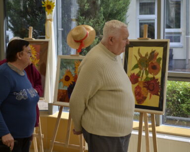 Zbliżenie. Troje seniorów ogląda wystawę obrazów. Na obrazach słoneczniki w kolorowych wazonach i tłach.