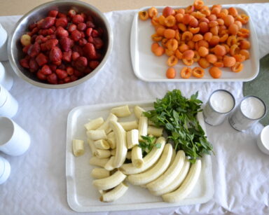 Zbliżenie. Stół, na nim, na tackach truskawki, morele, banany i gałązki mięty. Obok puszki mleka oraz białe filiżanki.