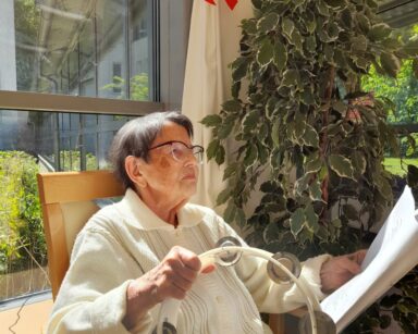 Zbliżenie. Seniorka w białym swetrze i koszulce siedzi przy oknie. W ręku trzyma tamburin i tekst piosenki na kartce.