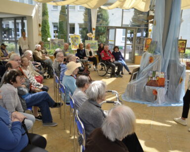 Sala. Przy kolumnie z woalem poustawiane są obrazy inspirowane Van Goghiem. Obok stoi kobieta i mówi do siedzących seniorów.