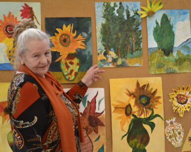Zbliżenie. Seniorka w kolorowej sukience pozuje przy tablicy z obrazami. Wskazuje na jeden obrazek z krajobrazem.