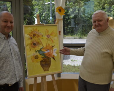 Zbliżenie. Dwóch mężczyzn pozuje do zdjęcia przy obrazie przedstawiającym słoneczniki w wazonie na żółtym tle.
