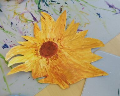 Zbliżenie. Na sole wycięty z kartonu i wymalowany na żółto i brązowo kwiat słonecznika. Pod pracą poplamiona farbami podkładka.
