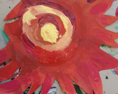 Zbliżenie. Na poplamionej kolorowymi farbami podkładce, leży czerwono-żółty wycięty z kartonu kwiat słonecznika.