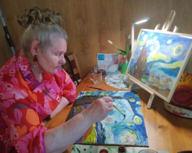 Zbliżenie. Seniorka siedzi przy stoliku z lamką, trzyma w ręku pędzel. Obok dwa podobne obrazy, 1 seniorka maluje farbami.