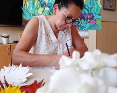 Zbliżenie. Kobieta w okularach i jasnej bluzce siedzi przy stole. Przed nią wycięte kwiaty słonecznika. W tle obraz irysów.