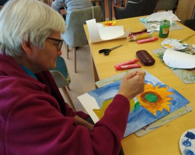 Sala. Seniorka maluje farbami obraz. Na kartce niebieskie tło, kwiat słonecznika i brązowy dzbanek. Na stole farby, woda.