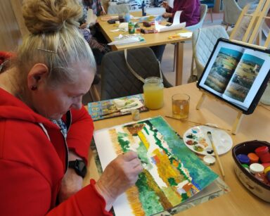 Sala. Seniorka maluje farbami obraz. Przed nią na sztaludze książka z obrazem, który kobieta kopiuje.