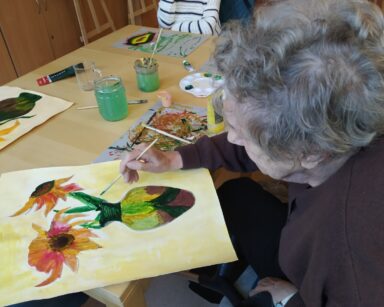 Zbliżenie. Przy stole pochylona seniorka. Maluje obraz na żółtym tle, kwiaty słonecznika w kolorowym wazonie.