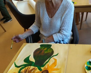 Sala. Seniorka w jasnym swetrze trzyma w ręku pędzel, patrzy na obraz na stole. Żółte tło, słoneczniki w zielonym wazonie.