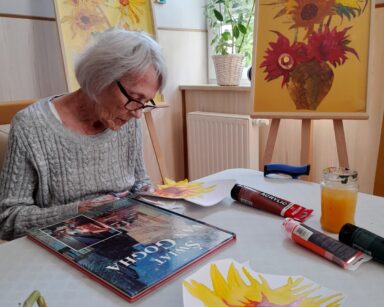 Sala. Seniorka w okularach i szarym swetrze siedzi przy stole. Maluje słonecznik. Przed nią książka pt. Świat Van Gogha.