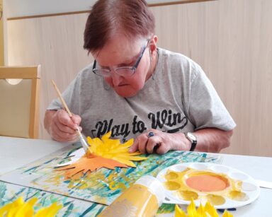 Zbliżenie. Seniorka przy stole maluje kwiat słonecznika. W ręku trzyma pędzel umazany pomarańczową farbą.