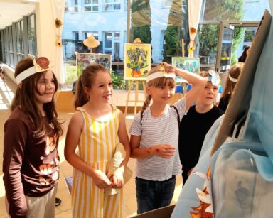 Sala. Czworo dzieci uśmiecha się i ogląda wystawę obrazów. W tle obrazy słoneczników, irysów oraz widok na drzewa.