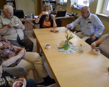 Sala. Grupa seniorów siedzi przy stole. Wszyscy jedzą z filiżanek łyżkami lody. W tle laptop, okna, regał z książkami.