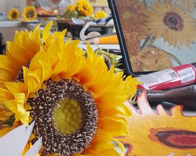 Zbliżenie. Na stole leży sztuczny kwiat słonecznika, obok tablet ze zdjęciem słonecznika, farby. W tle seniorka przy stole.