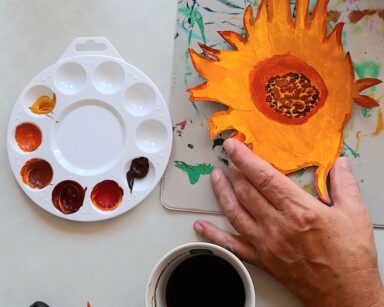 Zbliżenie. Stół. Wycięty kwiat słonecznika pomalowany farbą na żółto i brązowo. Farby. Kubek z kawą.