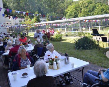Ogród. Przy stołach seniorzy. Nad stołami wiszą kolorowe proporczyki. W tle wolontariusze. Obok kobieta przy mikrofonie.